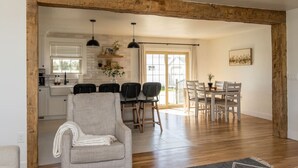 Un salón espacioso con techo de madera, una zona de comedor con sillas blancas y una mesa, y una cocina con armarios blancos y una ventana.