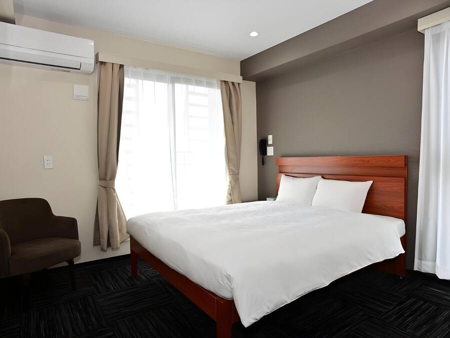 A hotel room with a bed, a chair, an air conditioning unit, and a telephone.