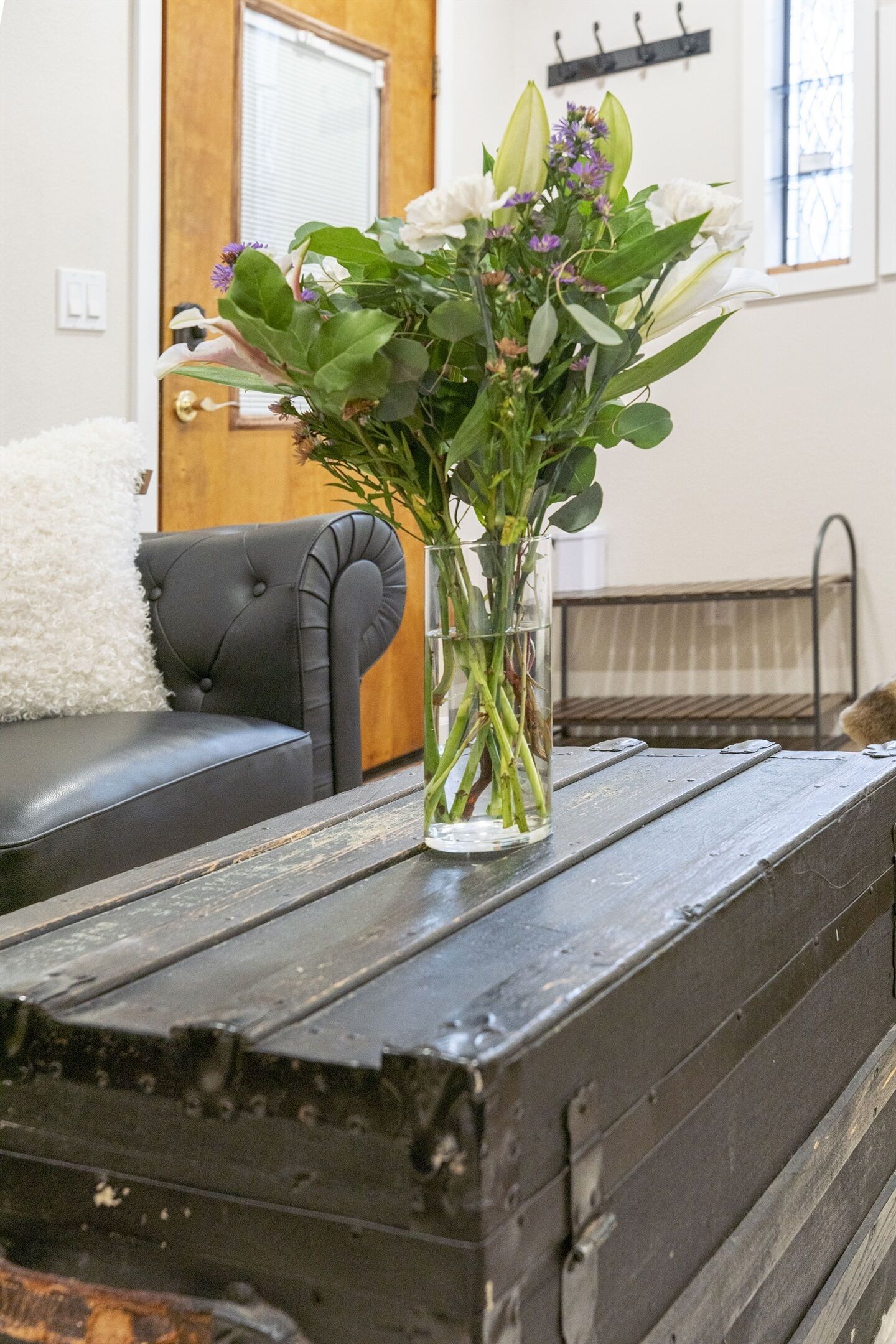A clear glass vase with flowers on a rustic wooden table in a room with a black leather sofa and a wooden door in the background.
