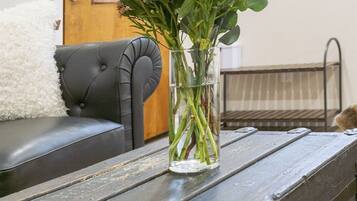 A clear glass vase with flowers on a rustic wooden table in a room with a black leather sofa and a wooden door in the background.