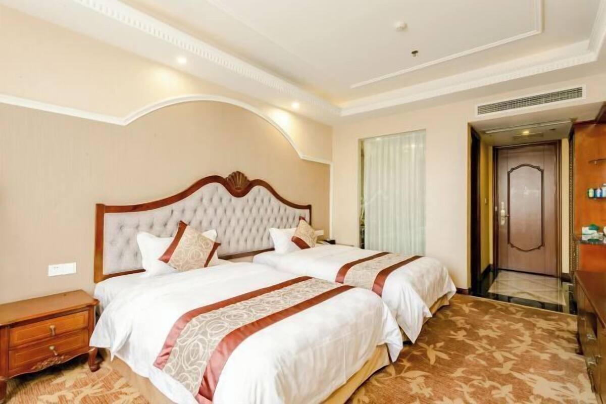 A hotel room with two beds, a wooden nightstand, a wardrobe, and a door leading to a bathroom.