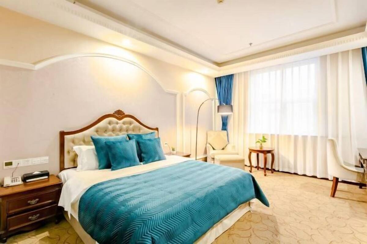 A hotel room with a large bed, a nightstand, a lamp, a chair, and a window with curtains.