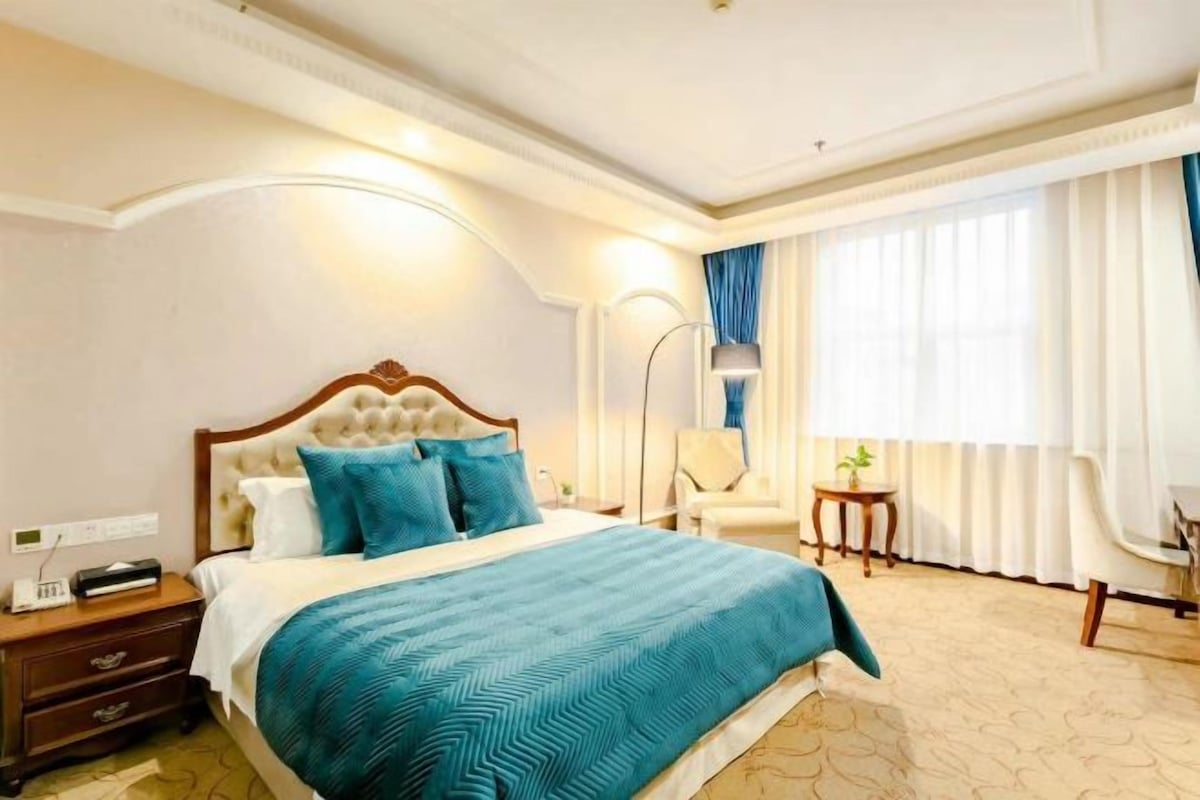 Una habitación de hotel con una cama grande, mesitas de noche, una silla, una pequeña mesa y un ventanal con cortinas.