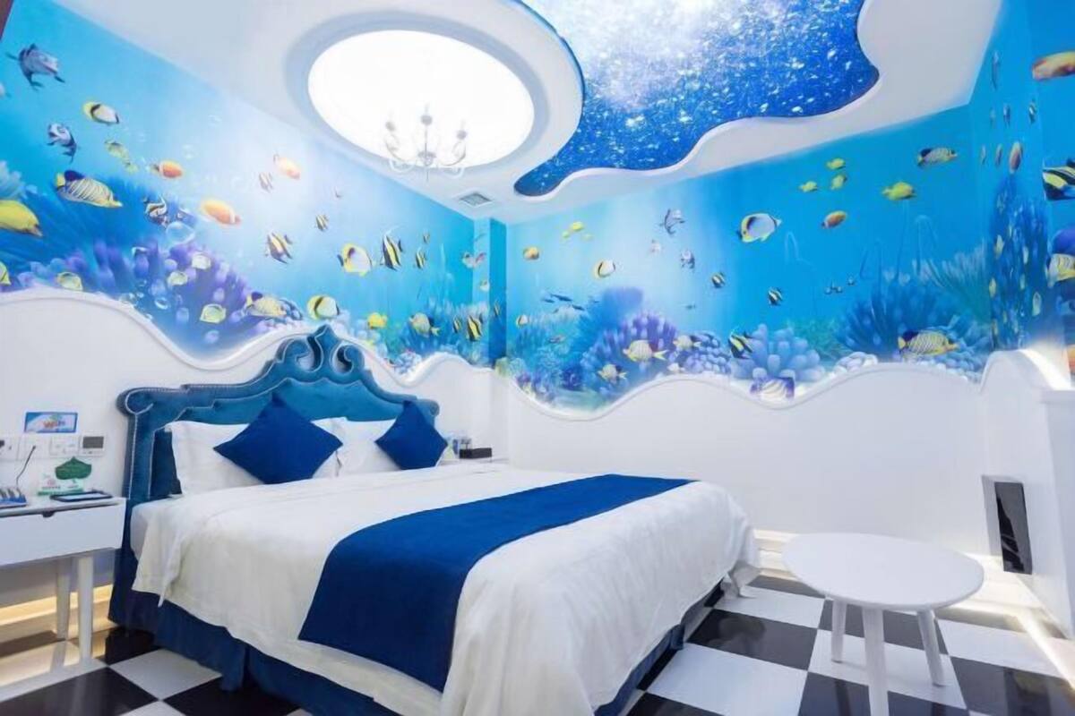 A bedroom with an ocean-themed mural, a bed with blue bedding, a round table, and a white nightstand.