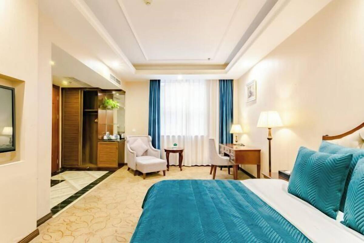 A hotel room with a large bed, a desk, a chair, a small table, and a window with curtains.