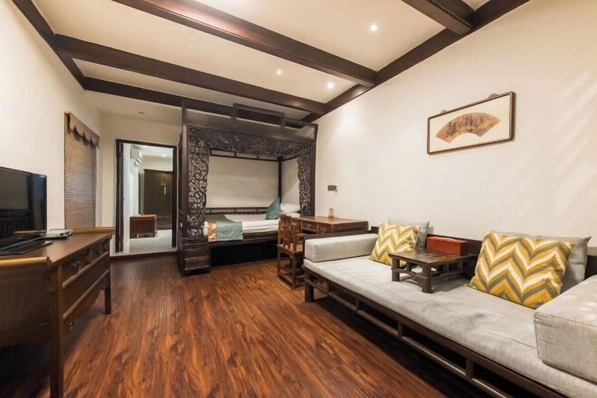 Um quarto com piso de madeira, um sofá com almofadas estampadas, uma escrivaninha, uma cama com cabeceira e um quadro pendurado na parede.