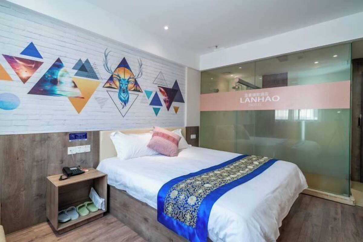 Ein Hotelzimmer mit Bett, Nachttisch, Hausschuhen, einer Wand mit Dekoration und einer Glastür zu einem Flur.