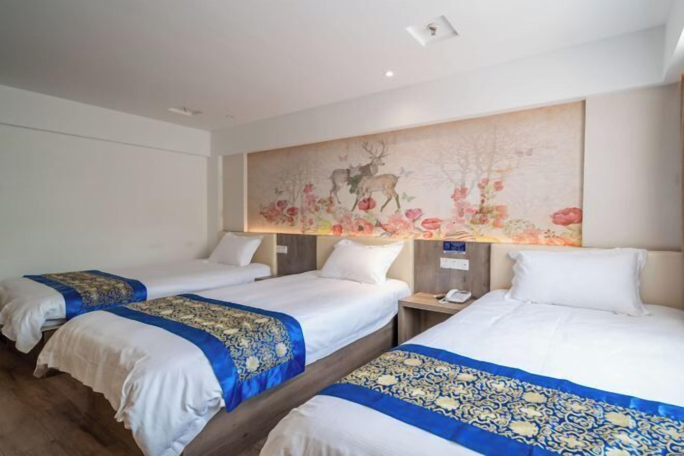 A hotel room with two beds, each with a blue and gold bedspread, white linens, and a large floral painting on the wall.