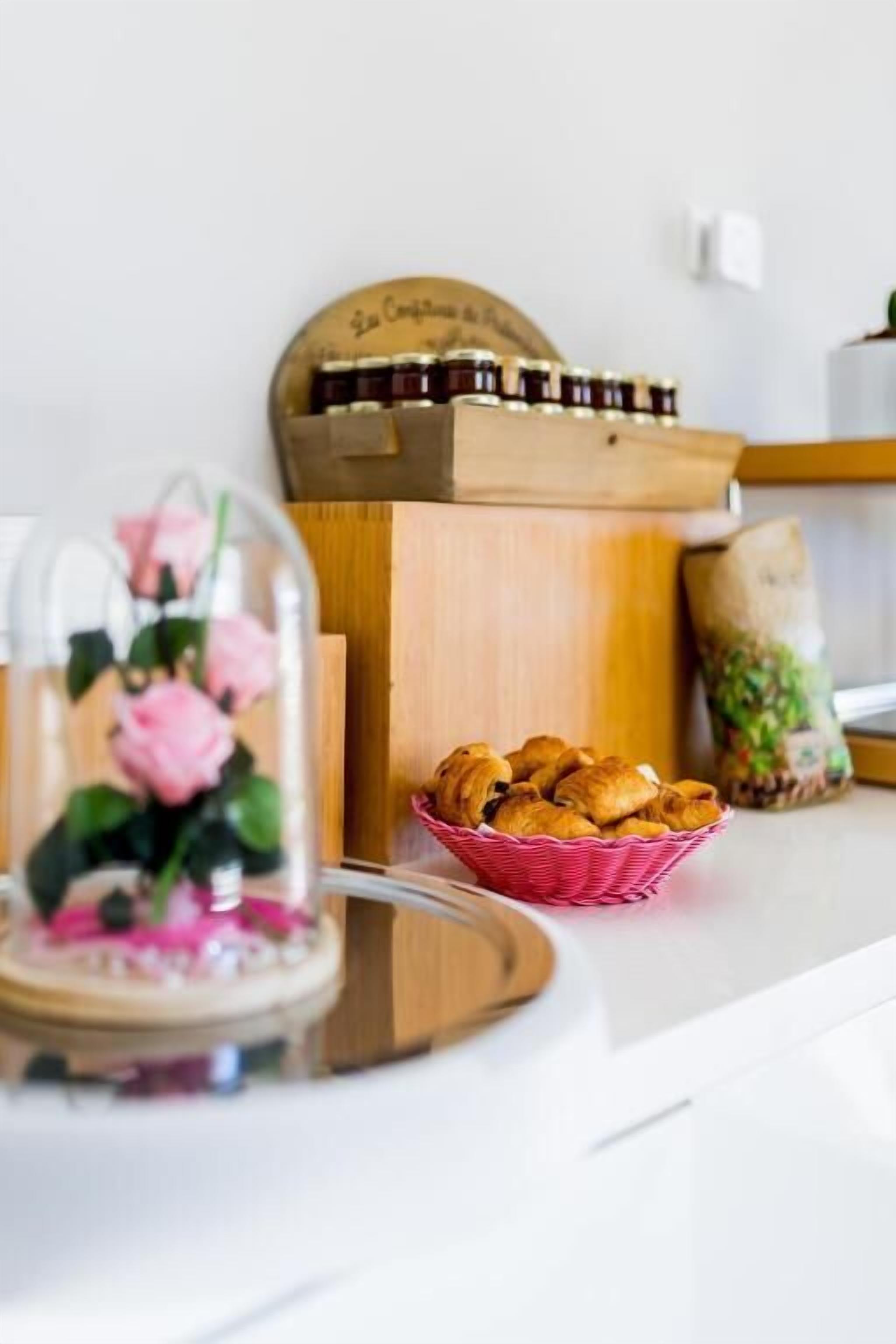 A kitchen counter with a glass dome containing pink roses, a basket of croissants, and a wooden box with jars.
