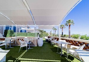 An outdoor patio with white chairs and tables, a covered area with a white canopy, and a view of palm trees and greenery.