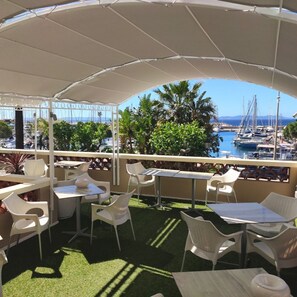 An outdoor terrace with white plastic chairs and tables, a white canopy, and a view of a marina with boats and palm trees.