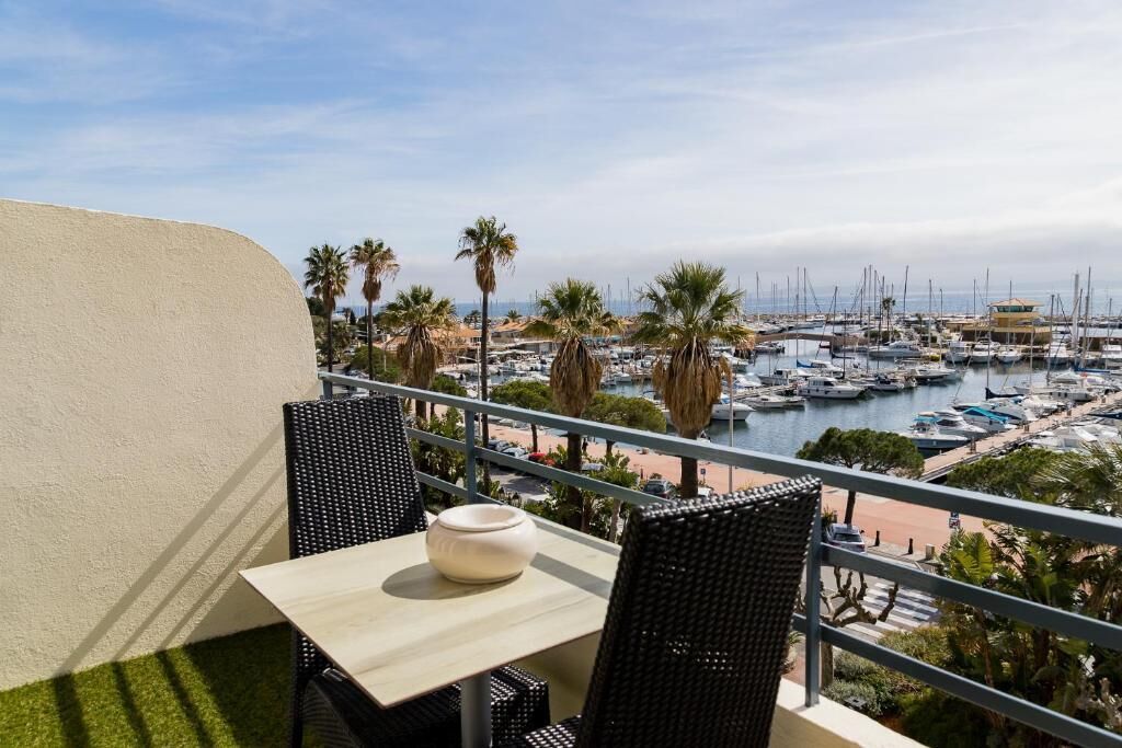 A balcony with a table and chairs overlooking a marina with boats and palm trees.