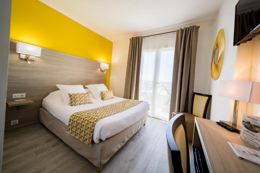 A hotel room with a bed, bedside table, lamp, and a view of the outside through the window.