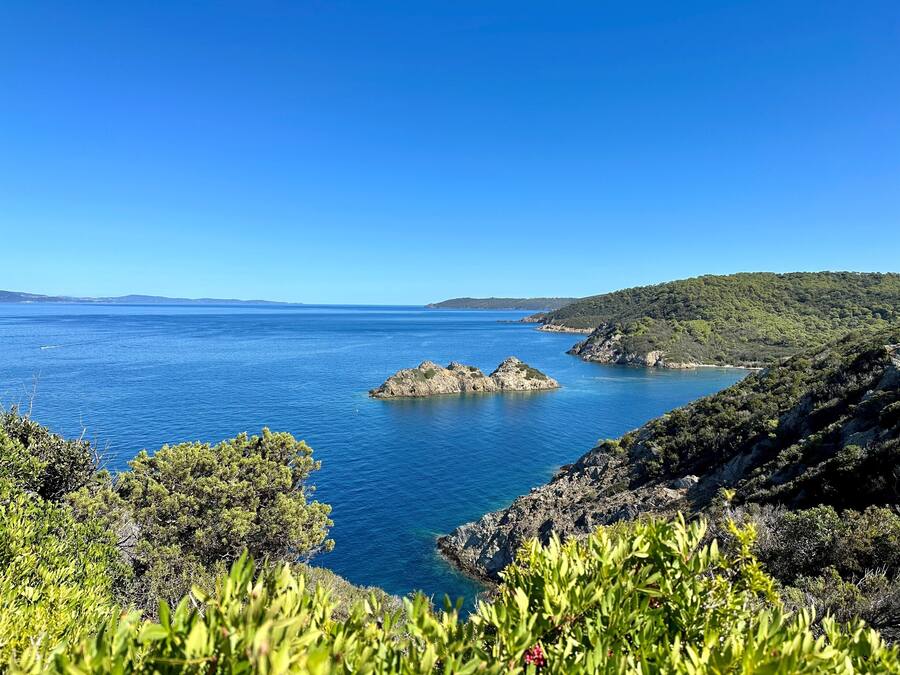 A coastal landscape with clear blue sea, rocky outcrops, and dense greenery.