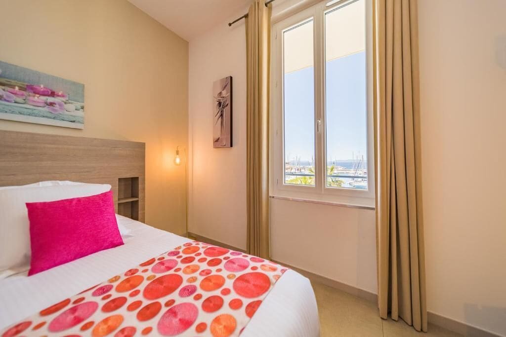 A hotel room with a bed, a window with curtains, a painting on the wall, and a city view.