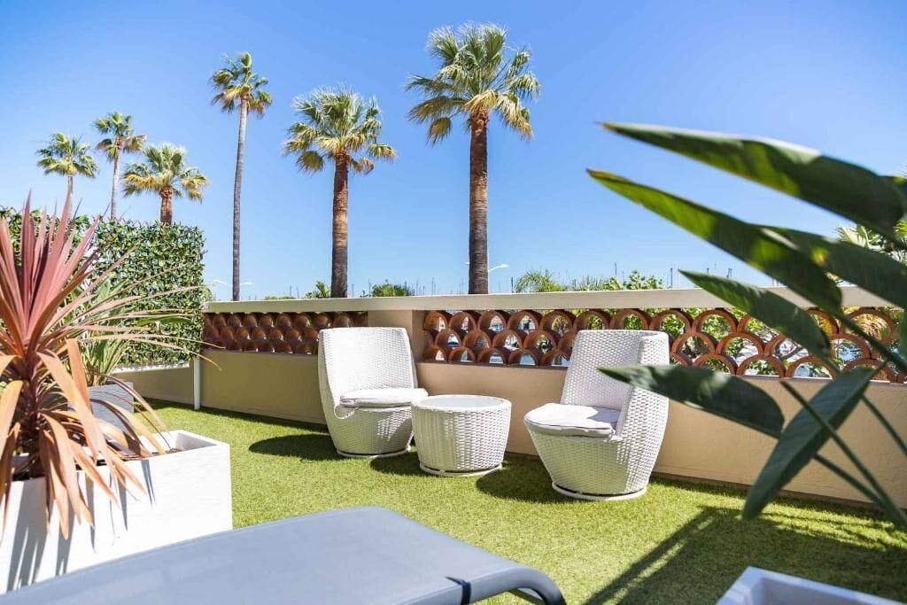 Un patio con mobiliario de mimbre blanco, palmeras y un muro decorativo.