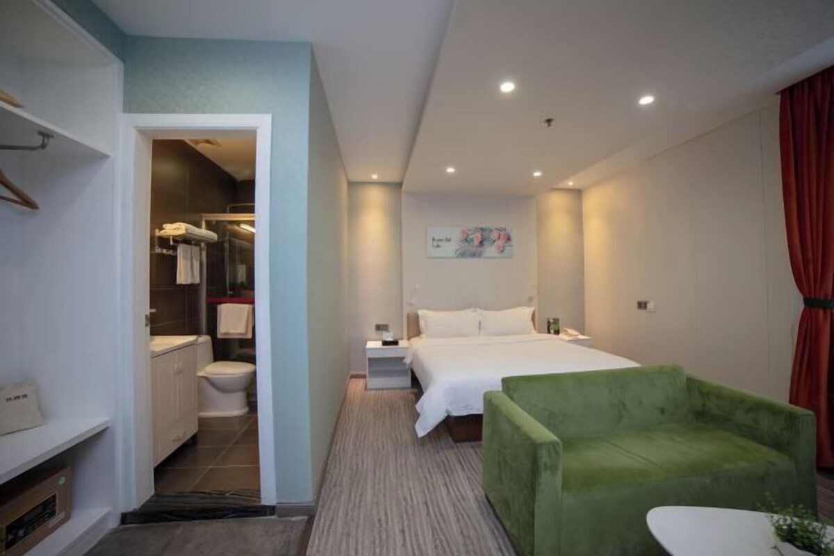 A hotel room with a bed, a green armchair, a small table, a painting on the wall, and a bathroom visible through an open door.