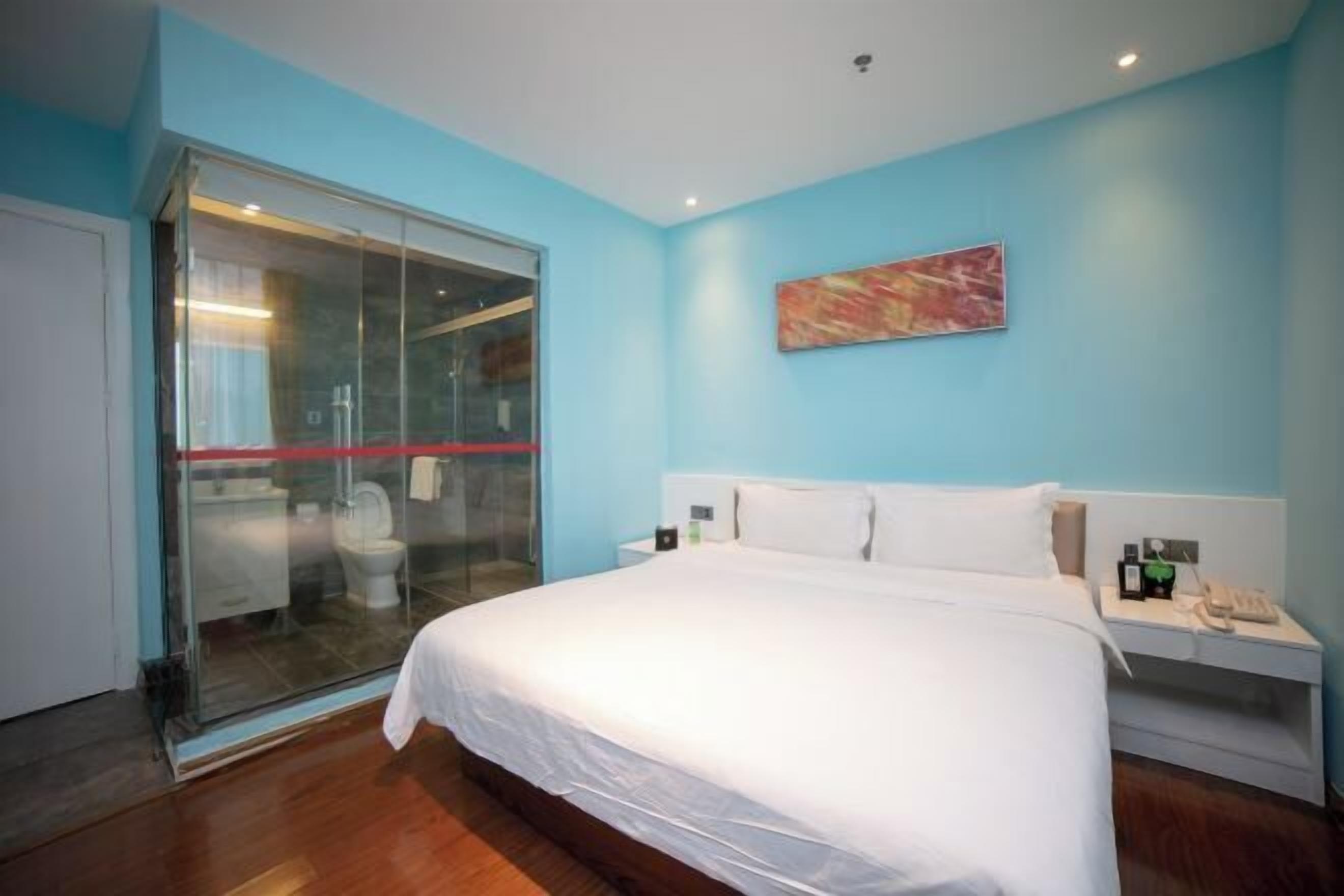 Quarto de hotel com uma cama grande, porta de vidro para um banheiro, mesa de cabeceira com telefone e um quadro na parede.