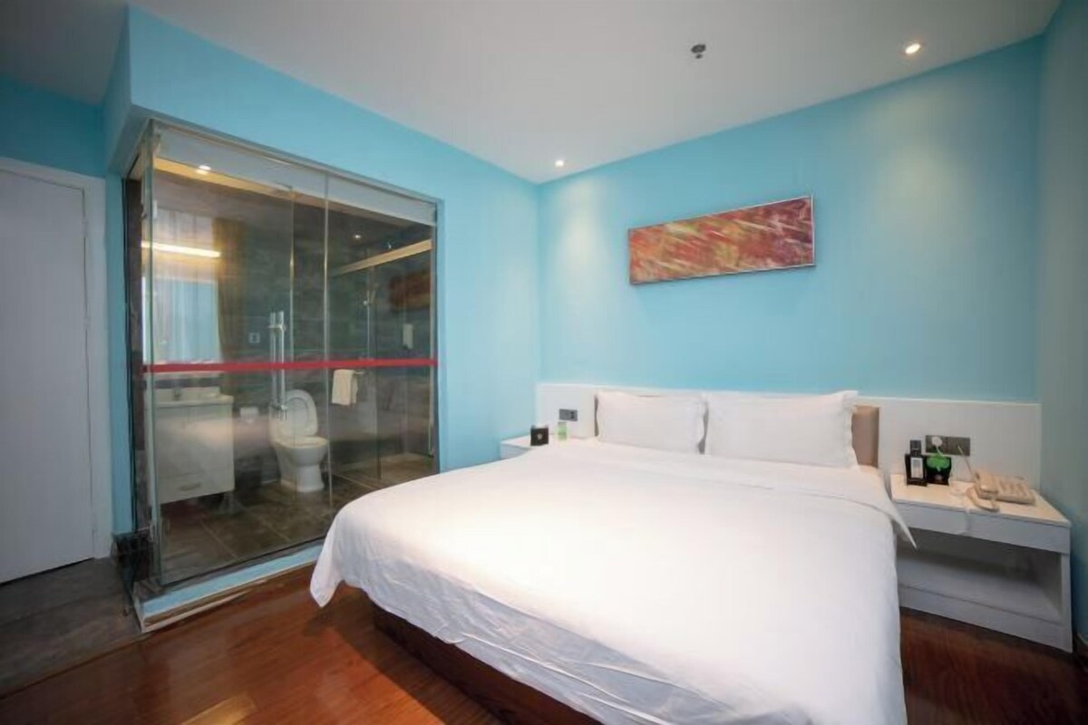 A hotel room with a large bed, a glass door leading to a bathroom, a nightstand with a phone, and a painting on the wall.