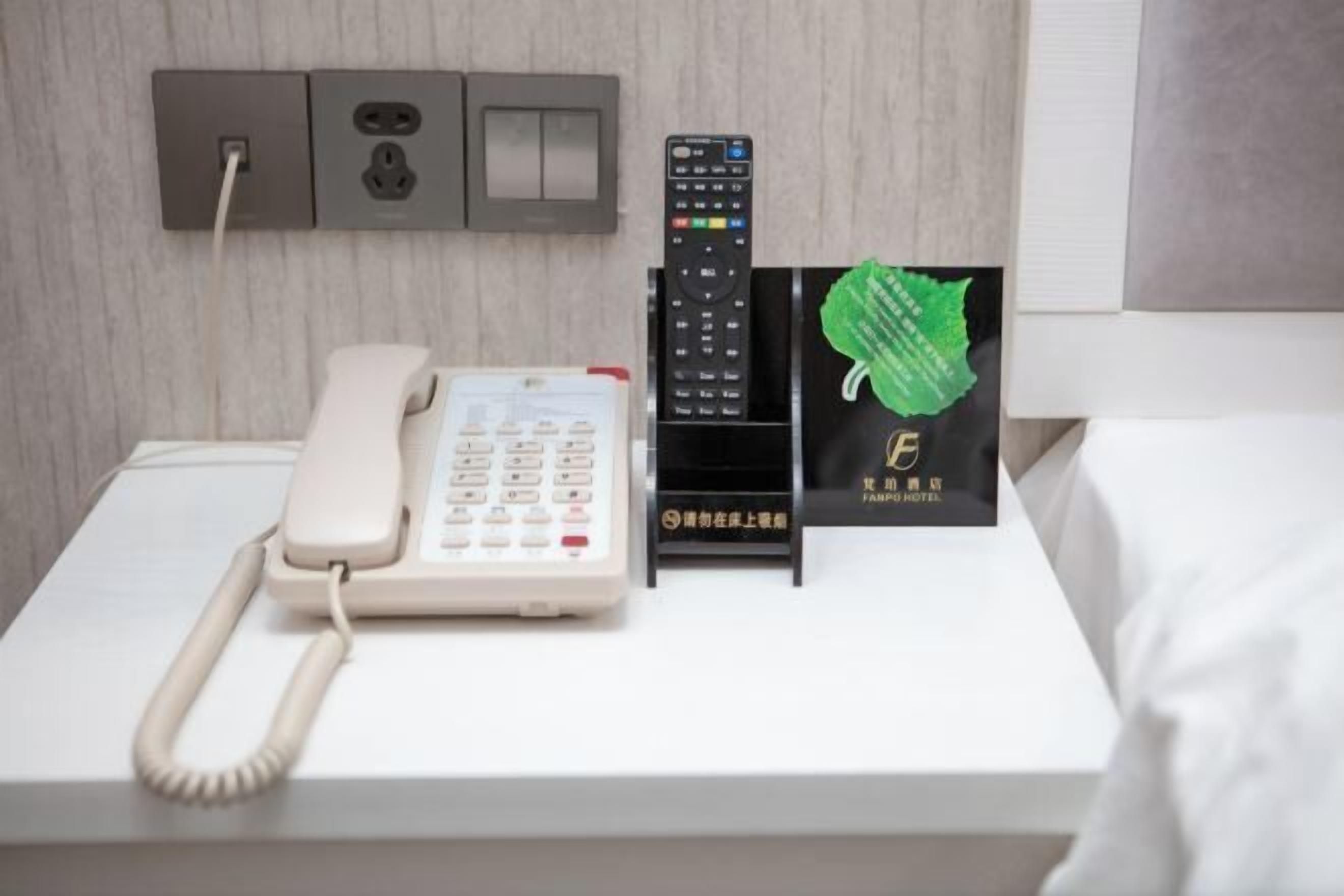 Habitación de hotel con una cama blanca, un teléfono blanco, un control remoto negro y un cartel de hotel verde.