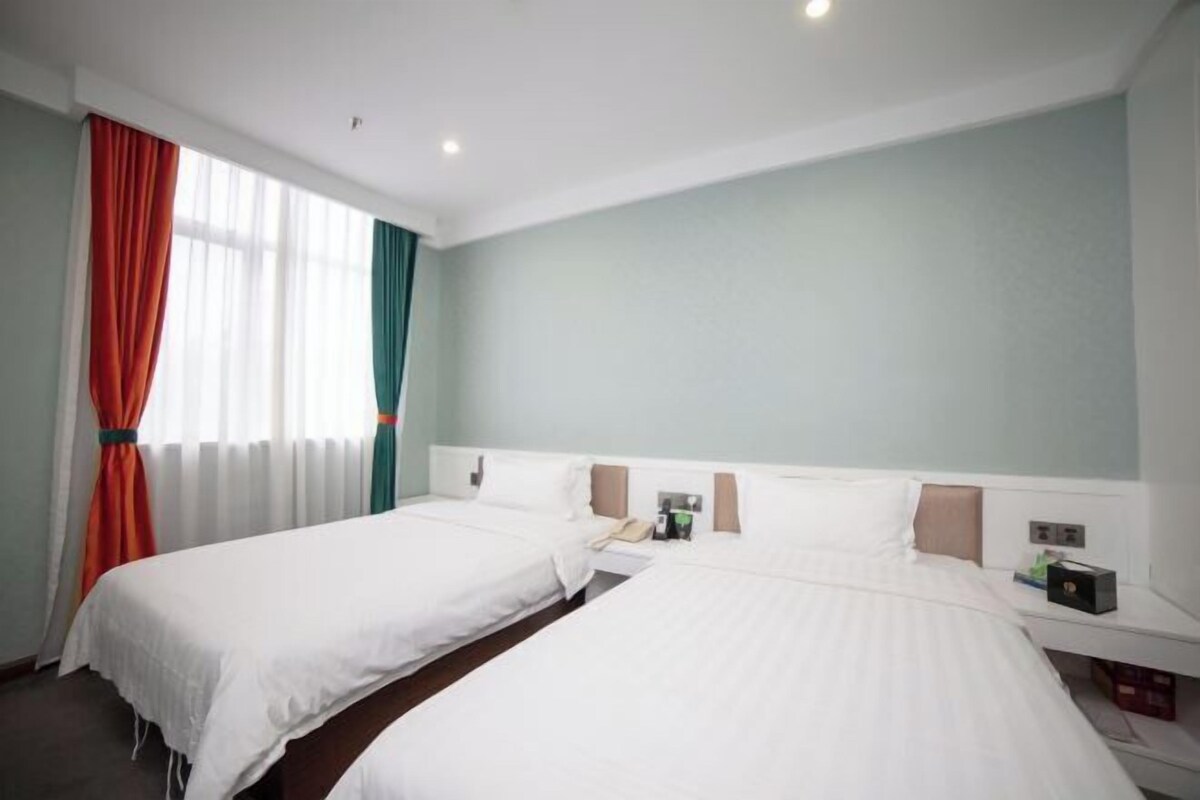 A hotel room with two beds, a window with curtains, a bedside table with a lamp, and a wall-mounted phone.
