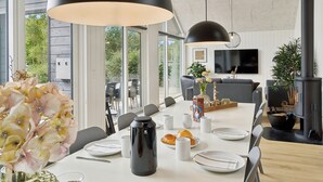 Un espace de restauration moderne, doté d’une grande table dressée pour quatre personnes, et entouré de larges fenêtres offrant une vue sur la verdure.