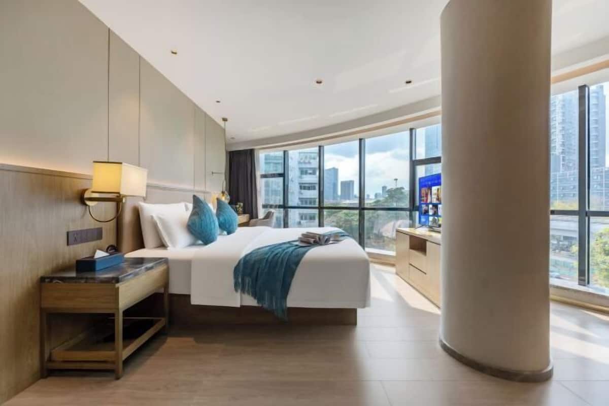 Habitación de hotel moderna con una cama grande, mesita de noche, lámpara y vistas a la ciudad a través de amplias ventanas.