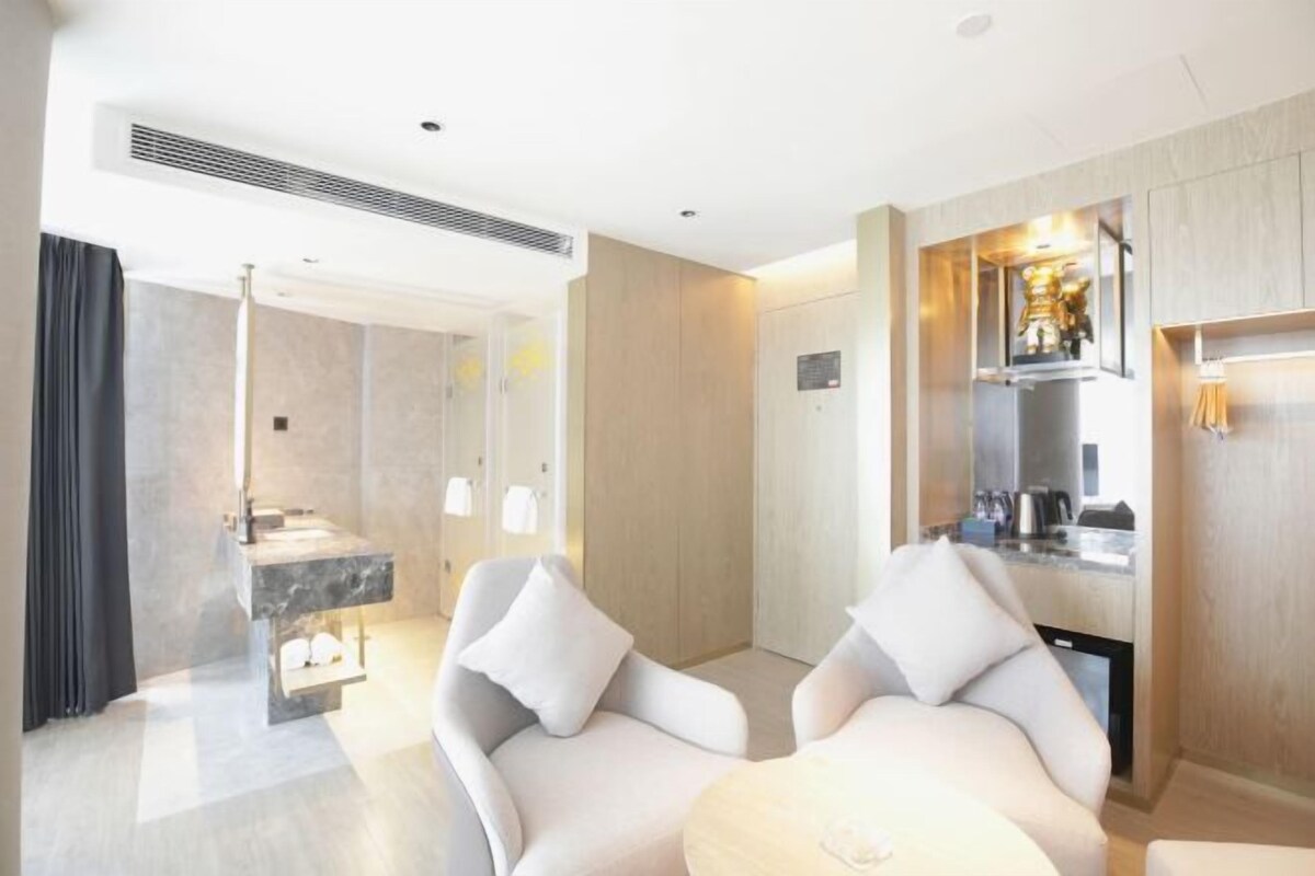 A modern hotel room with a marble bathroom, two white armchairs, a round table, and a built-in kitchen area.