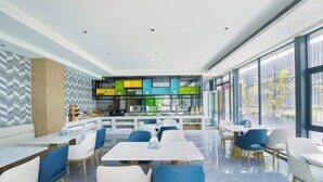 A modern dining area with white tables and blue chairs, a kitchen counter with colorful cabinets, and large windows offering a view outside. - Blueprint Cloud Hotel (Shenzhen Longcheng Plaza subway station store) (Shenzhen)