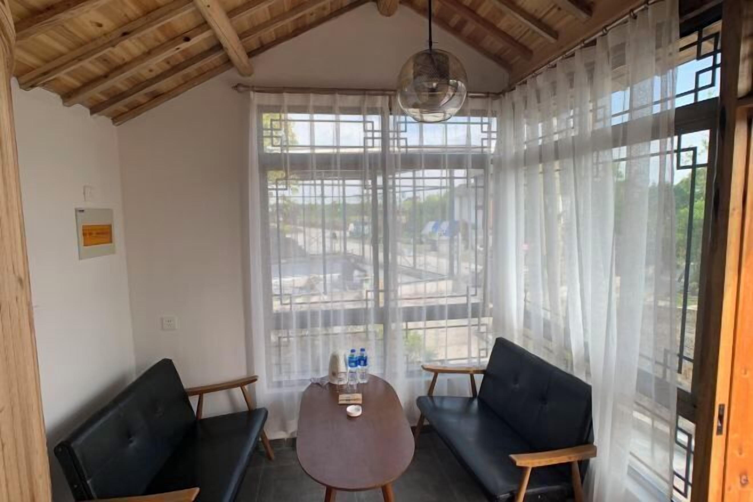 Quarto com teto de madeira, uma mesa redonda com duas cadeiras, um sofá e uma grande janela com cortinas leves.