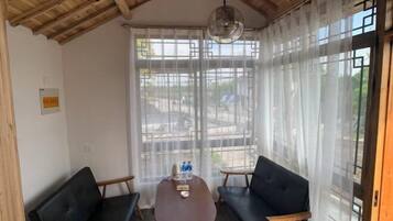 Quarto com teto de madeira, uma mesa redonda com duas cadeiras, um sofá e uma grande janela com cortinas leves.