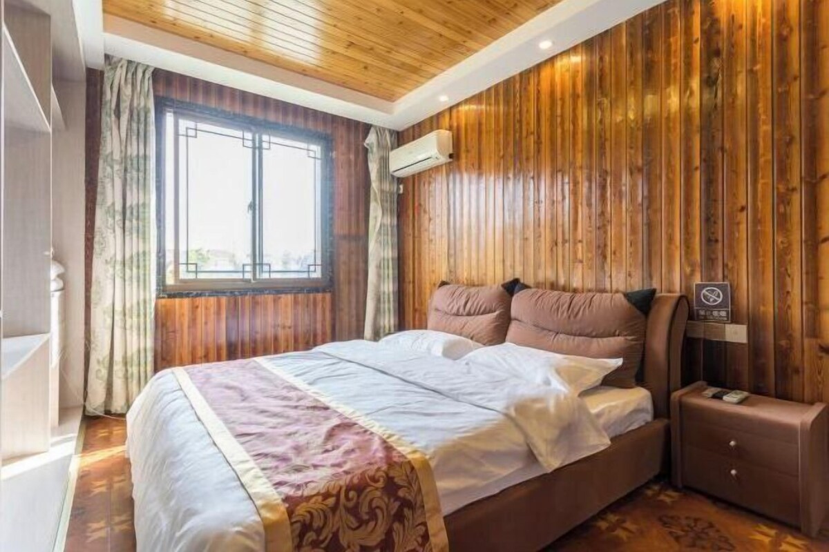 A bedroom with a wooden ceiling, a large window, a bed with white linens, and a nightstand.