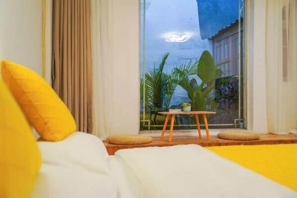 Une chambre à coucher avec un lit, des oreillers jaunes, une petite table avec une plante et une vue sur la verdure à l’extérieur.