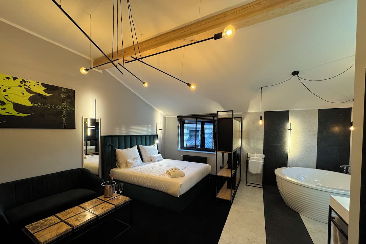 Ein modernes Hotelzimmer mit Bett, Sofa, einem kleinen Tisch mit einem Getränk, einem großen Fenster, einem Badezimmer mit Badewanne und einem Bild an der Wand.