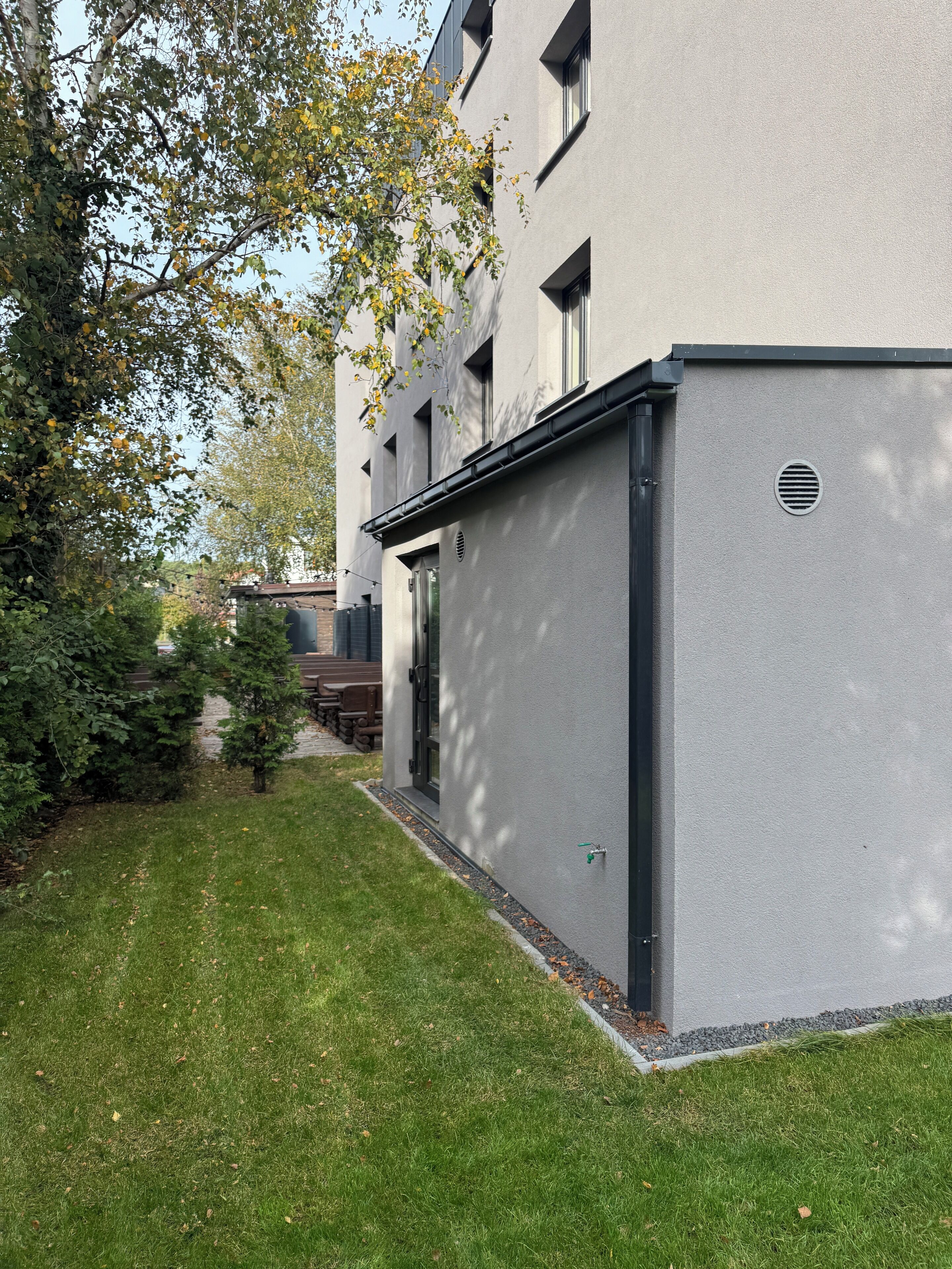 Un edificio moderno con fachada gris, un pequeño jardín con césped y árboles, y un patio con mobiliario exterior.