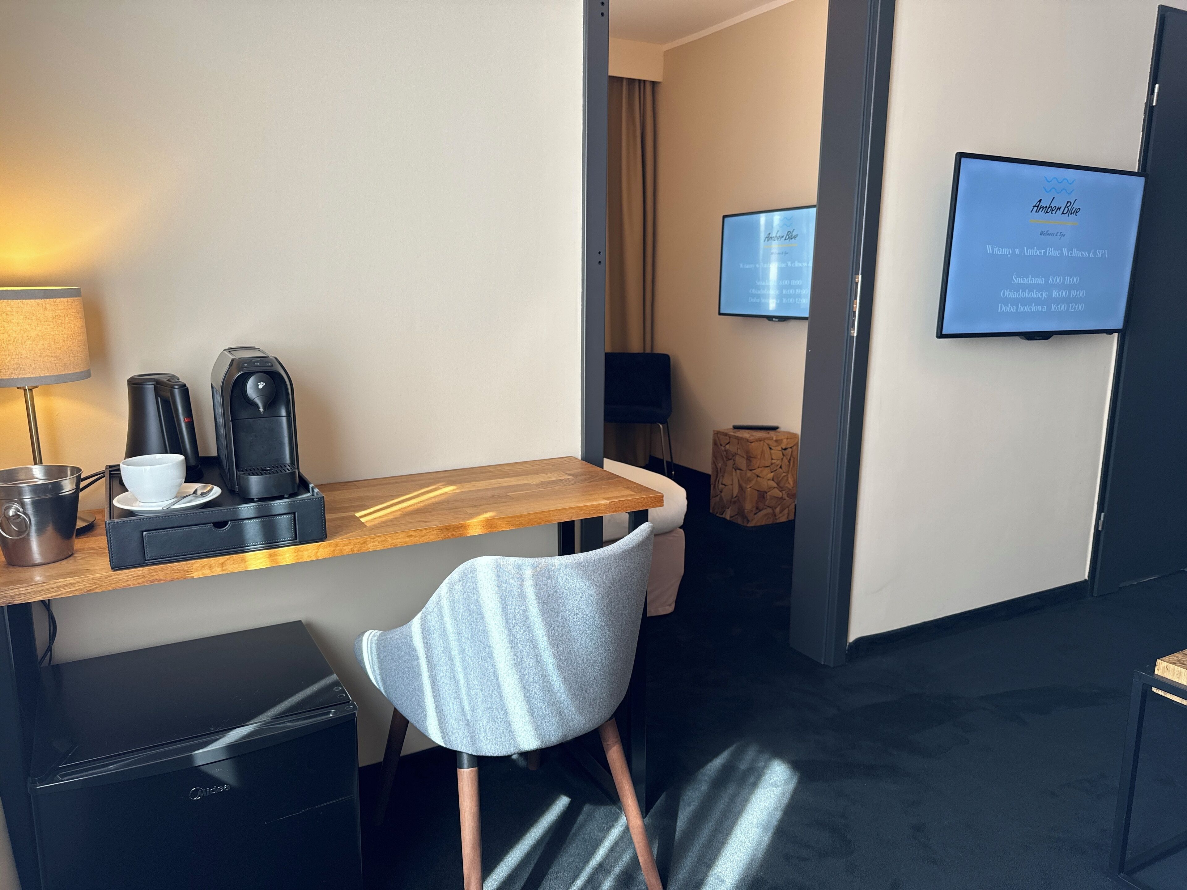 Habitación de hotel con un escritorio de madera, una silla, cafetera, taza, cuchara, mueble pequeño, televisor en la pared y lámpara.