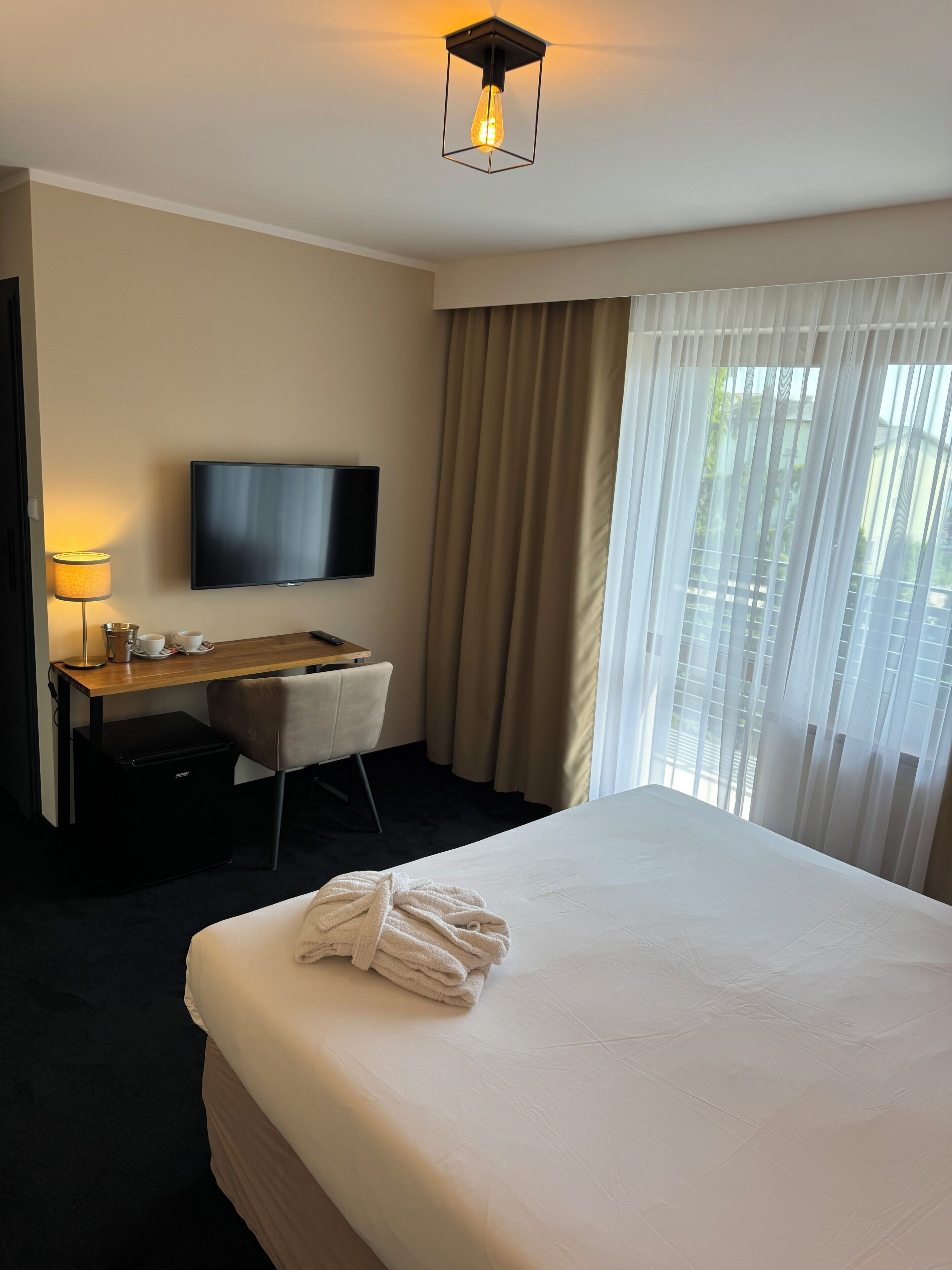 A hotel room with a bed, a desk with a chair, a television, and a window with curtains.