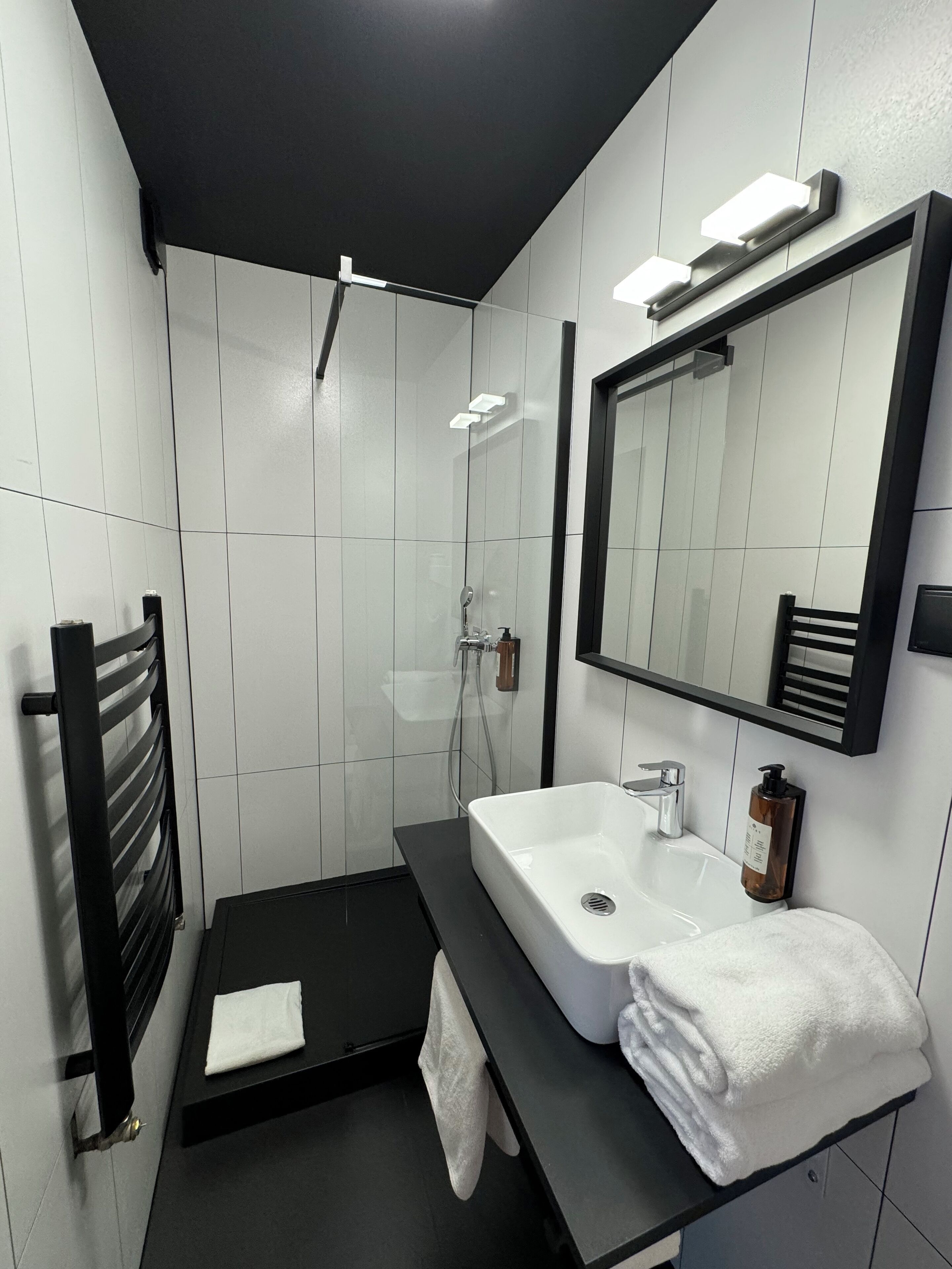 Un baño moderno con un lavamanos negro, azulejos blancos, una cabina de ducha de vidrio y un perchero para toallas.