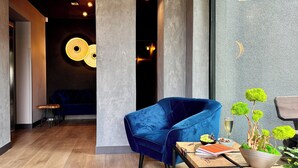 Un interior moderno con un sillón de terciopelo azul, una mesa de madera con libros y una planta, y una pared con luminarias circulares.
