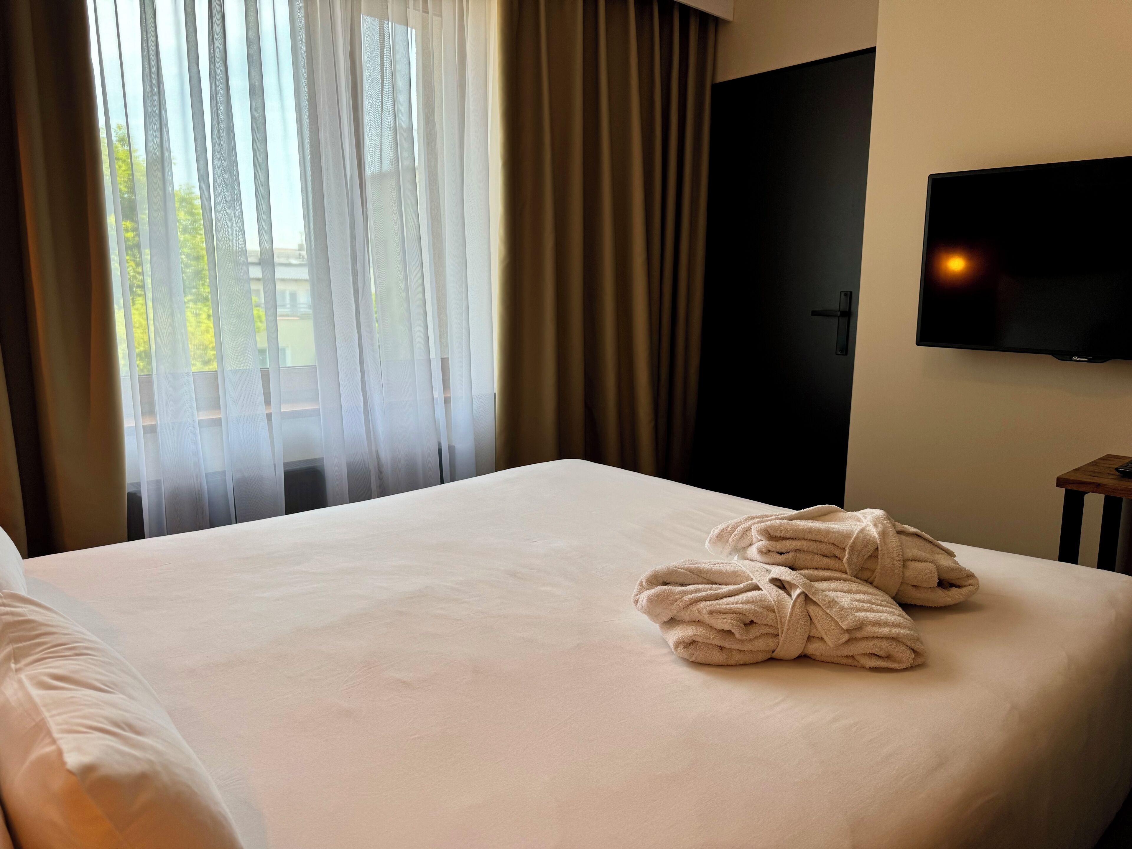 A hotel room with a bed, a TV mounted on the wall, and a window with sheer curtains.
