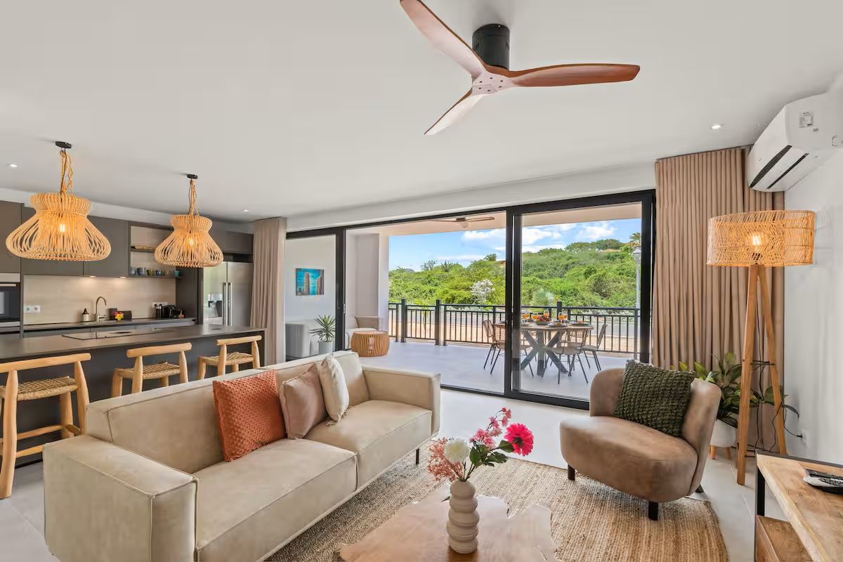 Una sala de estar moderna con sofá, sillón y una mesa de centro, una cocina con taburetes de bar y un balcón con mobiliario exterior.