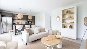 A modern living room with a sofa, coffee table, and dining area. There is a kitchen in the background with a refrigerator, stove, and dining chairs. - The Breeze (Sint Michiel)