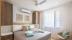 A modern hotel room with a large bed, a ceiling fan, an air conditioning unit, a window with sheer curtains, and a small seating area. - The Breeze (Sint Michiel)