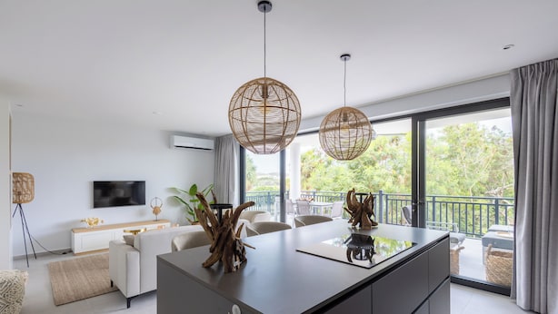 A modern living room with a central island, two hanging rattan light fixtures, a flat-screen TV, and a balcony with outdoor seating. - The Breeze (Sint Michiel)