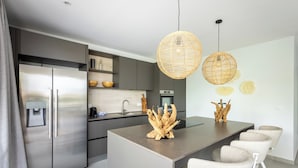 A modern kitchen with a stainless steel refrigerator, a built-in island with bar stools, and a hanging light fixture. - The Breeze (Sint Michiel)