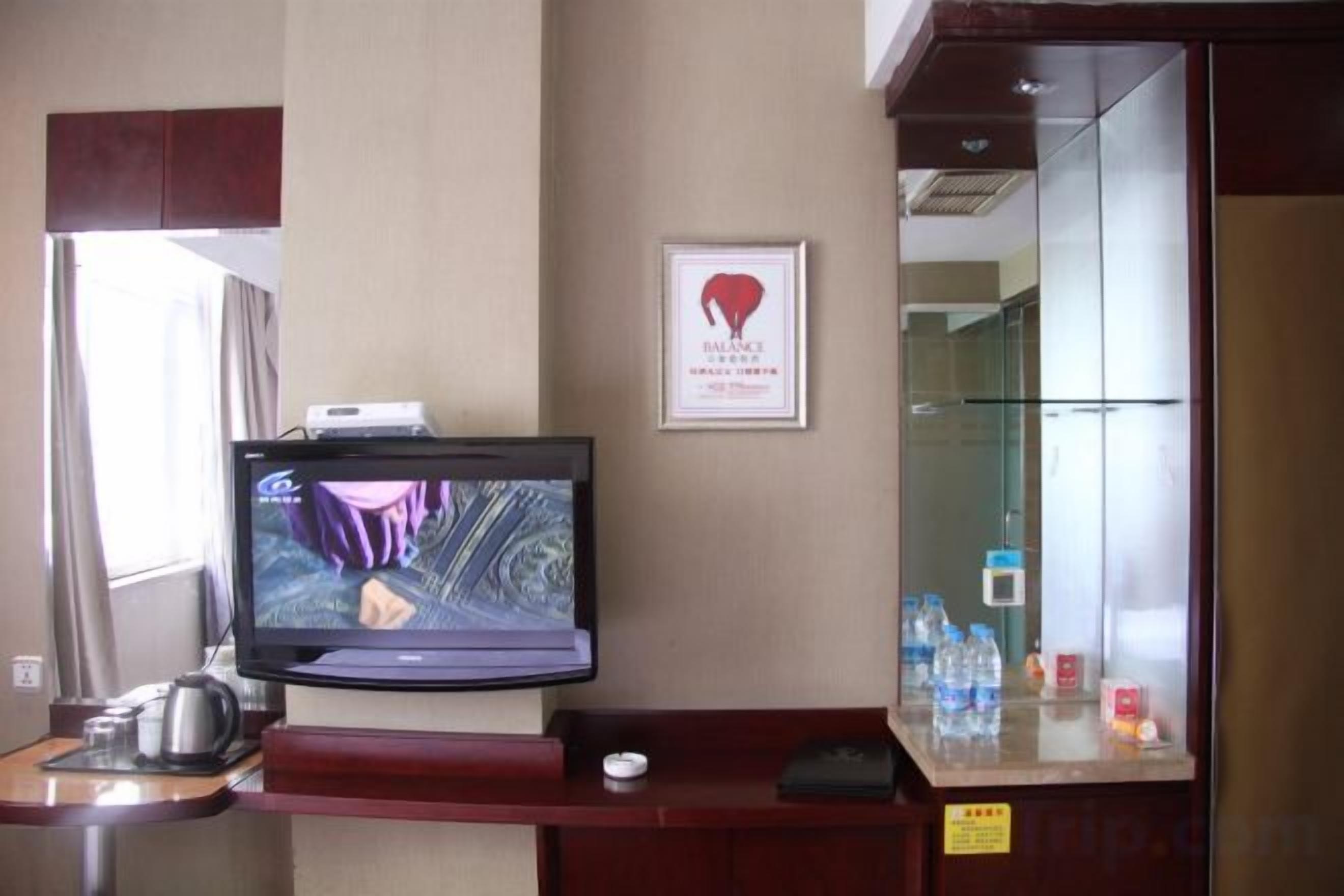 Zona de recepción de un hotel con un televisor, un certificado enmarcado en la pared y una vitrina de vidrio con botellas de agua.