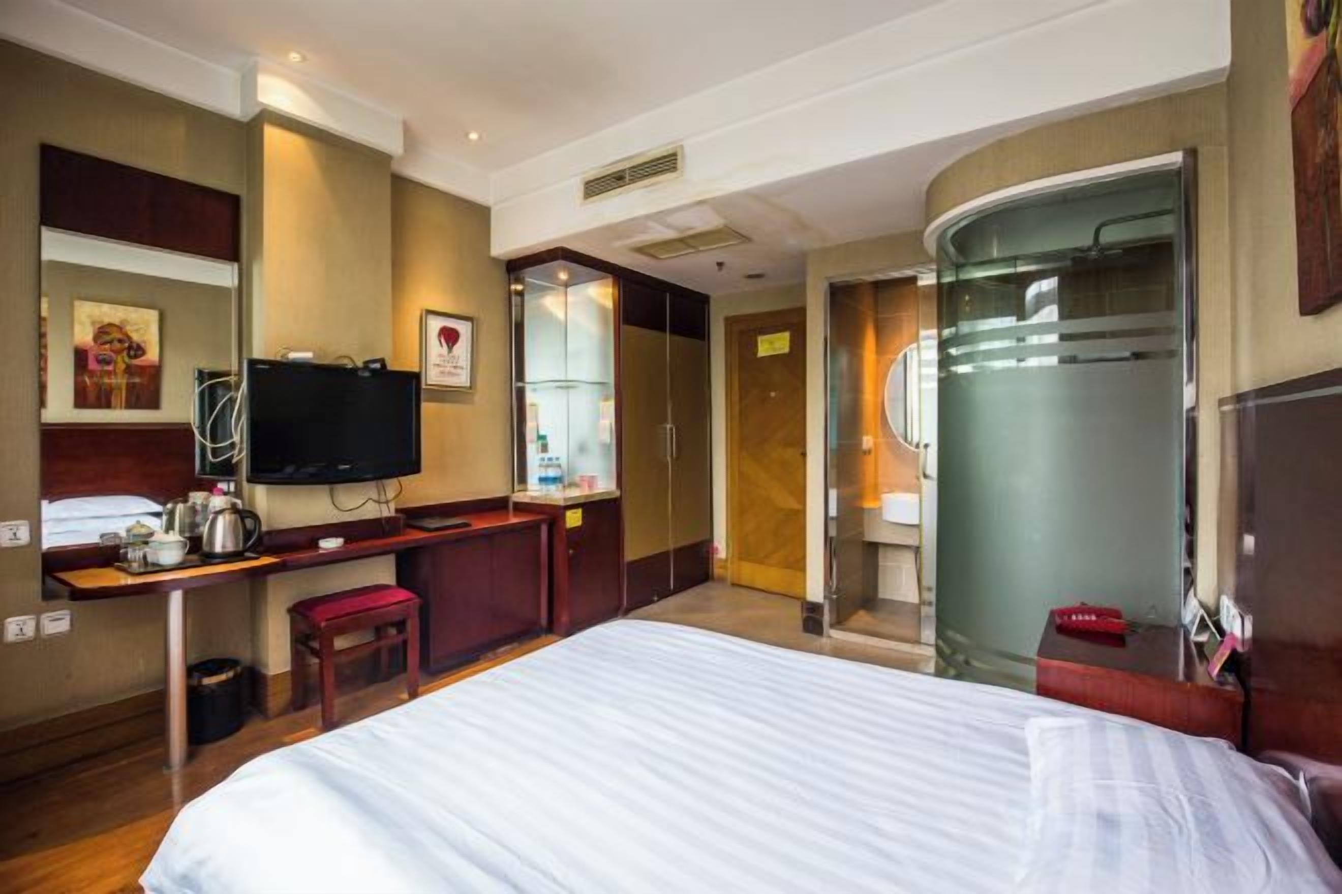Een hotelkamer met een bed, een bureau met een televisie, een spiegel en een badkamer met een douchecabine van glas.