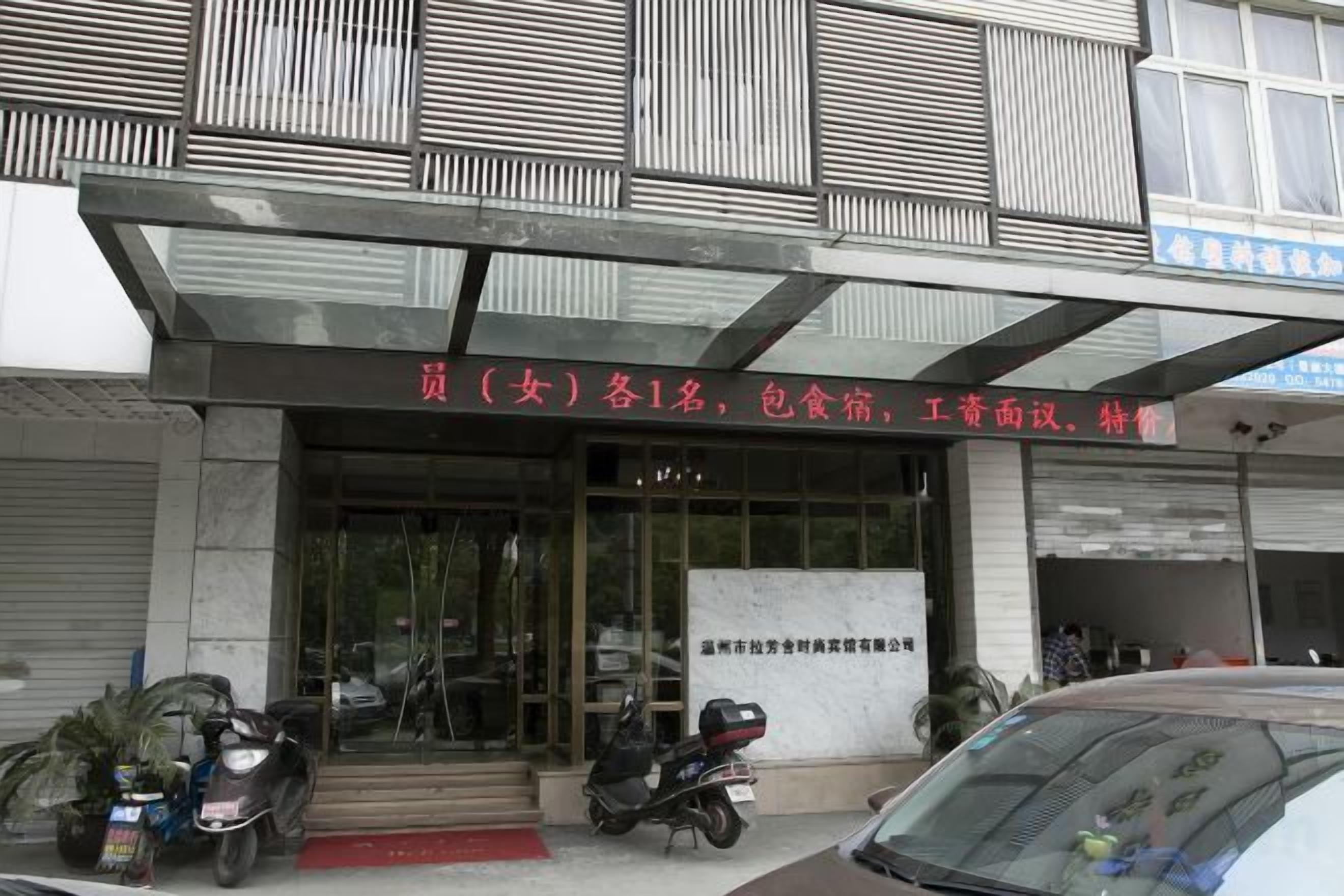 A building with a covered entrance, a signboard with text, and parked vehicles.