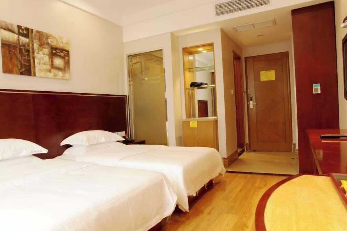 Habitación de hotel con dos camas, cabecera de madera, un escritorio, un espejo y una puerta que da al baño.