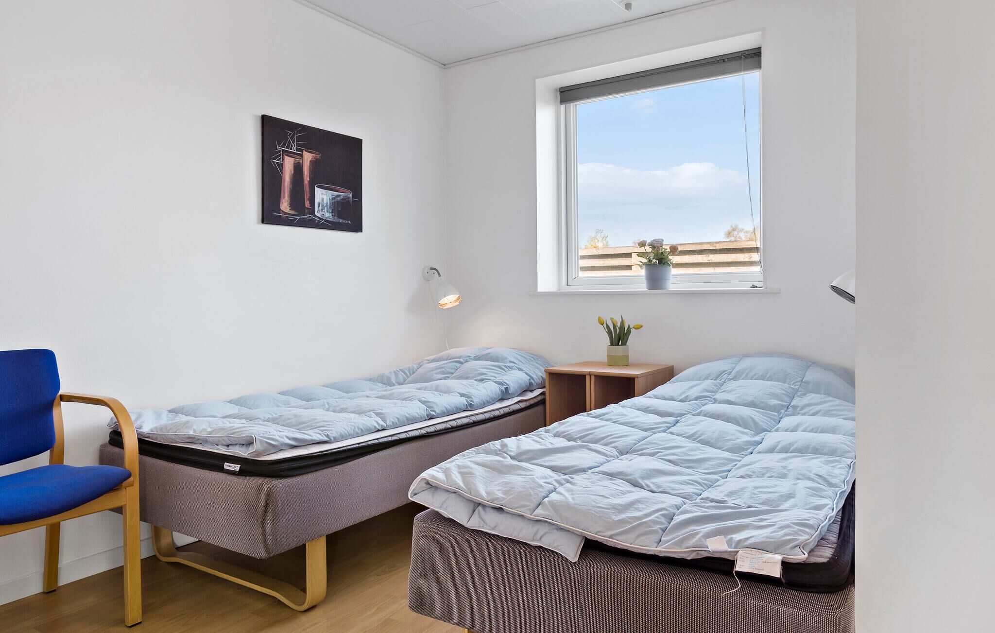 Une petite pièce bien éclairée, avec deux lits simples, une chaise, un bureau et une fenêtre donnant sur le ciel et une clôture.